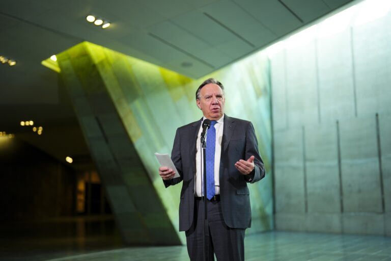 Quebec Premier François Legault in a suit speaking at a microphone in a modern building with angular architectural elements.
