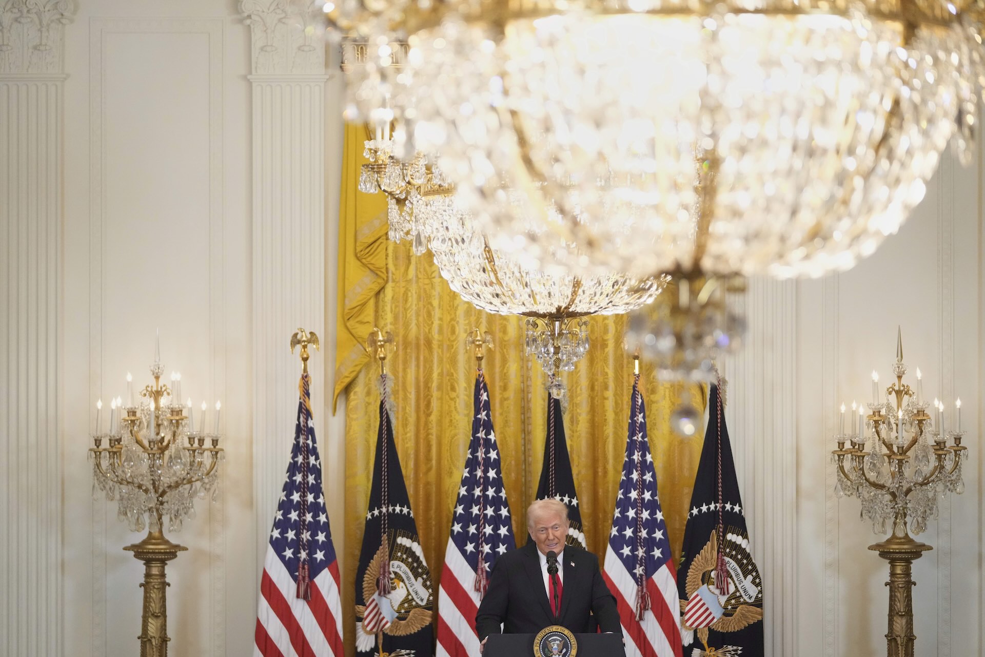 U.S. President Donald Trump standing at a podium with multiple U.S. flags behind him, under a large chandelier in an ornate room.