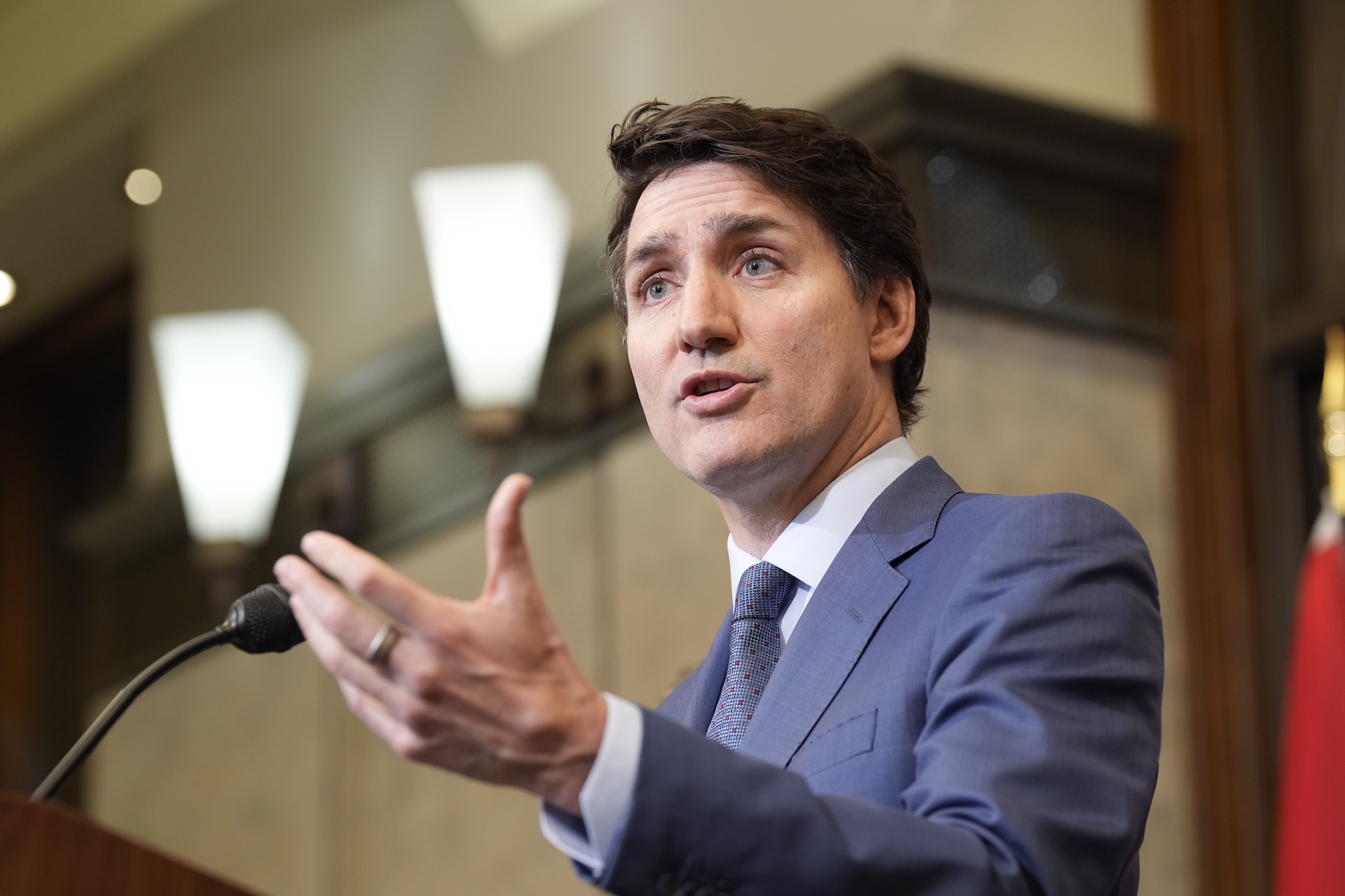 Prime Minister Justin Trudeau in a suit speaking at a podium and gesturing with his left hand, in a room with modern light fixtures.