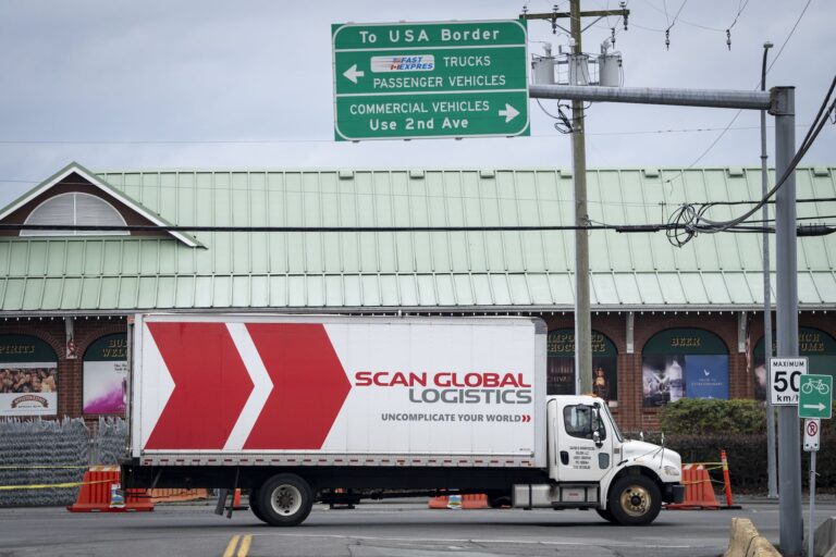 A truck with "Scan Global Logistics" branding on it near the U.S.-Canada border. A sign above it says "To USA Border" and has arrows for "Trucks Passenger Vehicles" and "Commercial Vehicles."