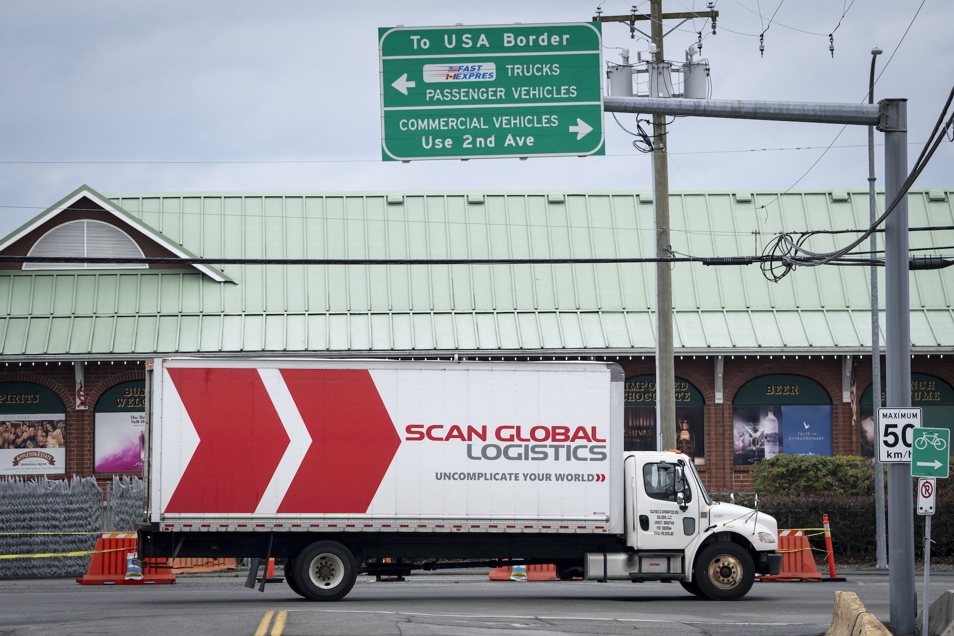 A truck with "Scan Global Logistics" branding on it near the U.S.-Canada border. A sign above it says "To USA Border" and has arrows for "Trucks Passenger Vehicles" and "Commercial Vehicles."