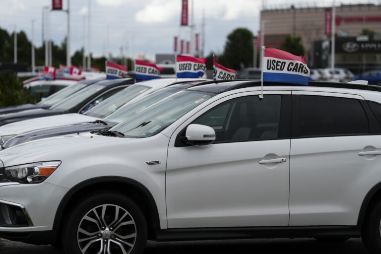 Row of parked vehicles with "Used Cars" flags on a dealership lot, with a white SUV in the front of the row.