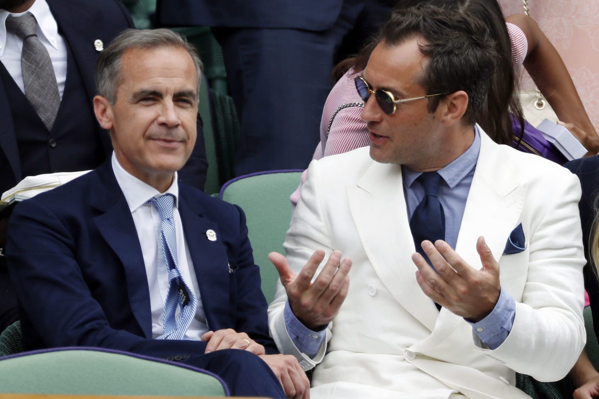 An image of Jude Law, wearing a white suit and sunglasses, in animated conversation with a smiling Mark Carney, who is wearing a dark suit.