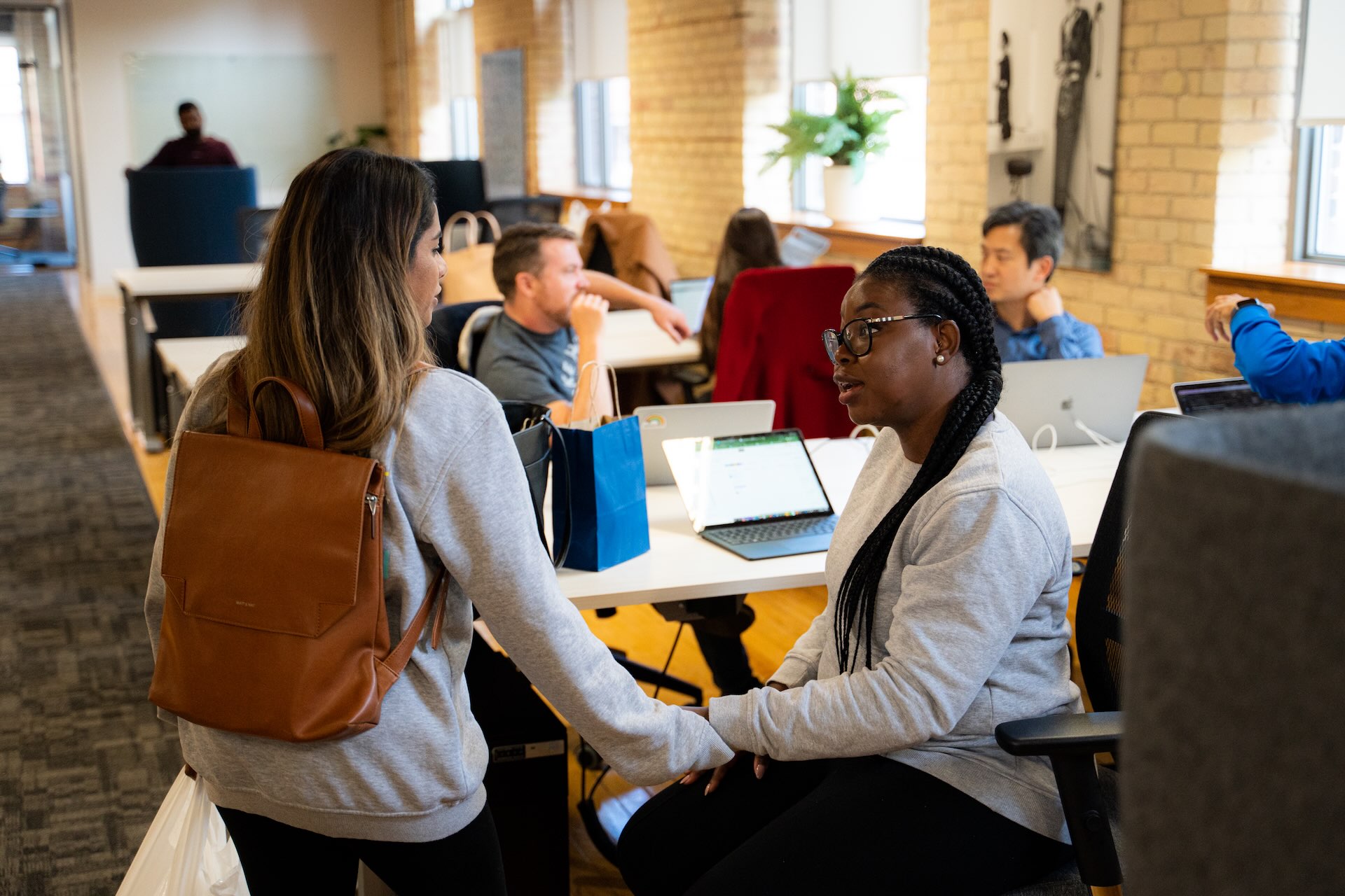 Two employees wearing grey sweaters talk in the foreground, while other employees work on laptops on tables behind them.