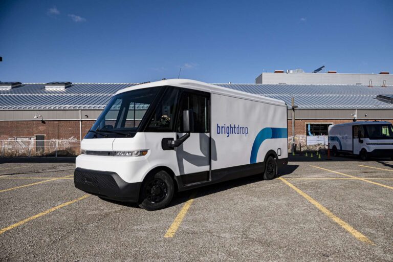 A shot of a large white utility van in a parking lot in front of a warehouse-style building. The van has a blue stripe and "BrightDrop" written on the side.