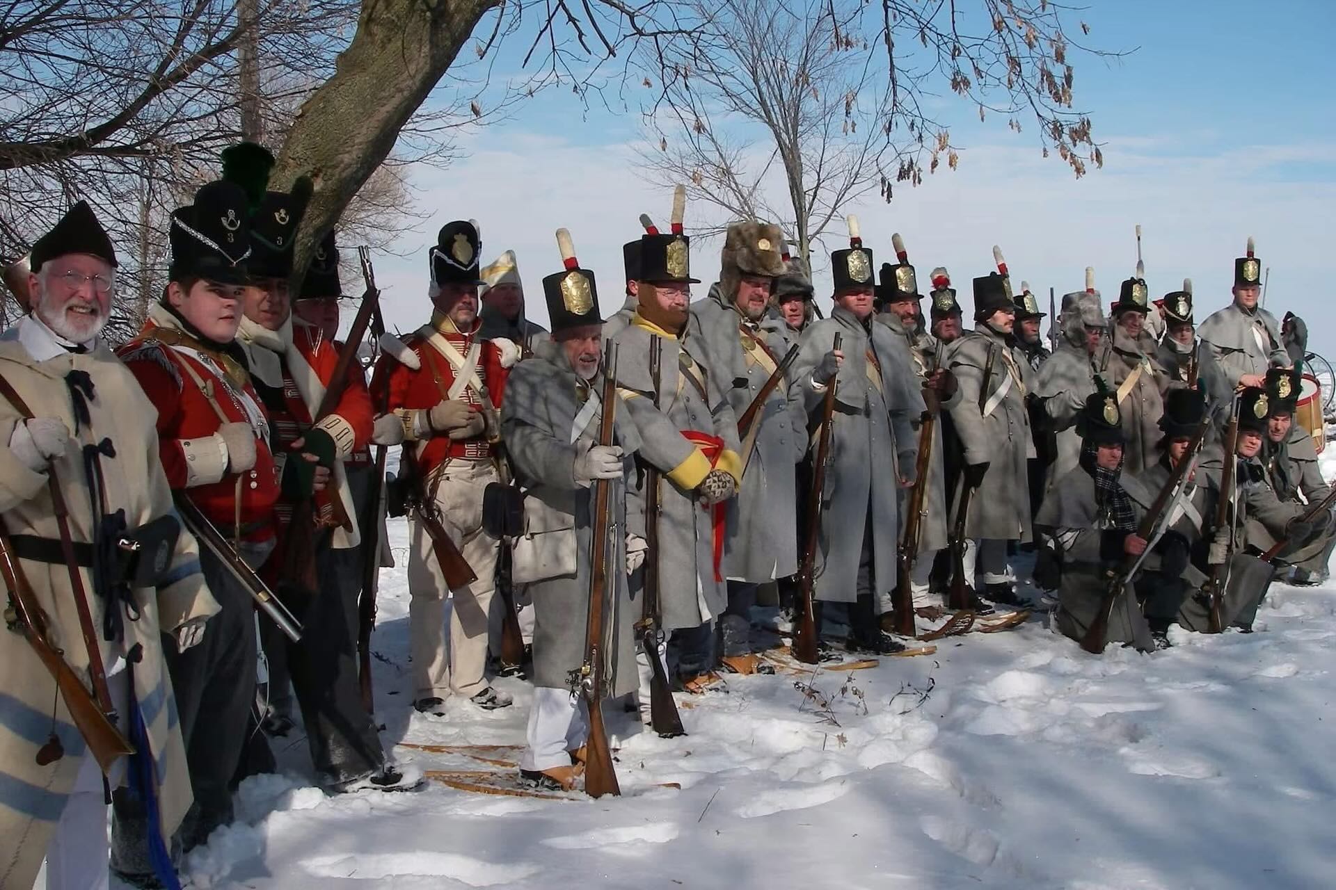 A line of more than 20 men standing or kneeling in the snow, dressed for a War of 1812 re-enactment of the Battle of Ogdensburg. Some wear red coats and others are in grey. Most are holding rifles.