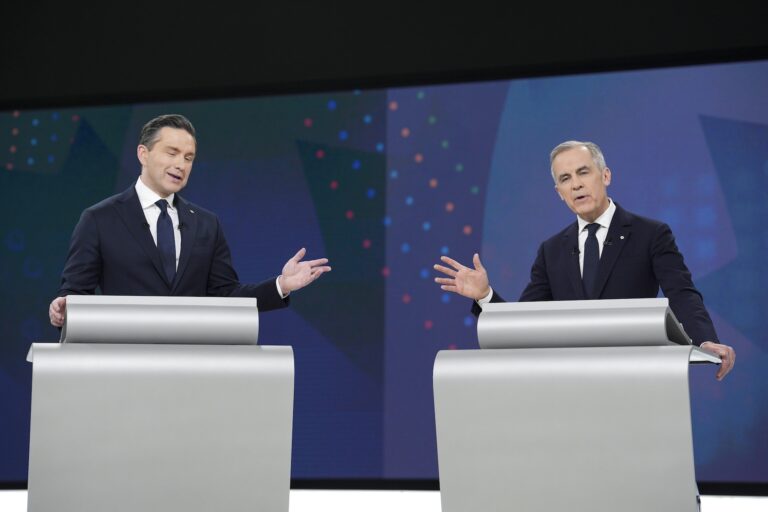 A debate shot showing Pierre Poilievre and Mark Carney side by side at silver-grey lecterns. Both are gesturing toward the other, with one hand slightly raised, while speaking.