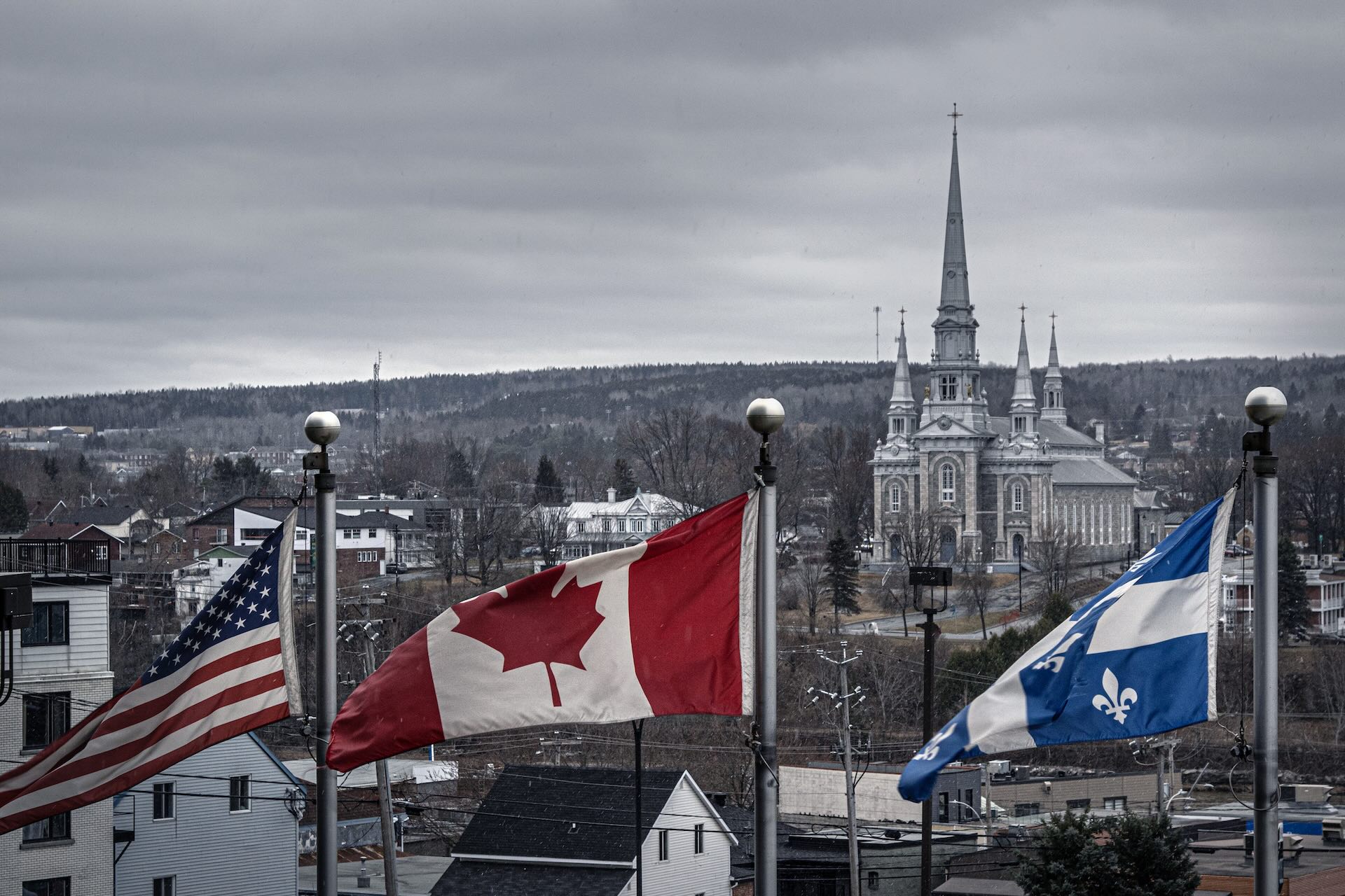 Three flags—the United States, Canada, and Quebec—fly in front of the church of St-Georges building on an overcast day in a small town.