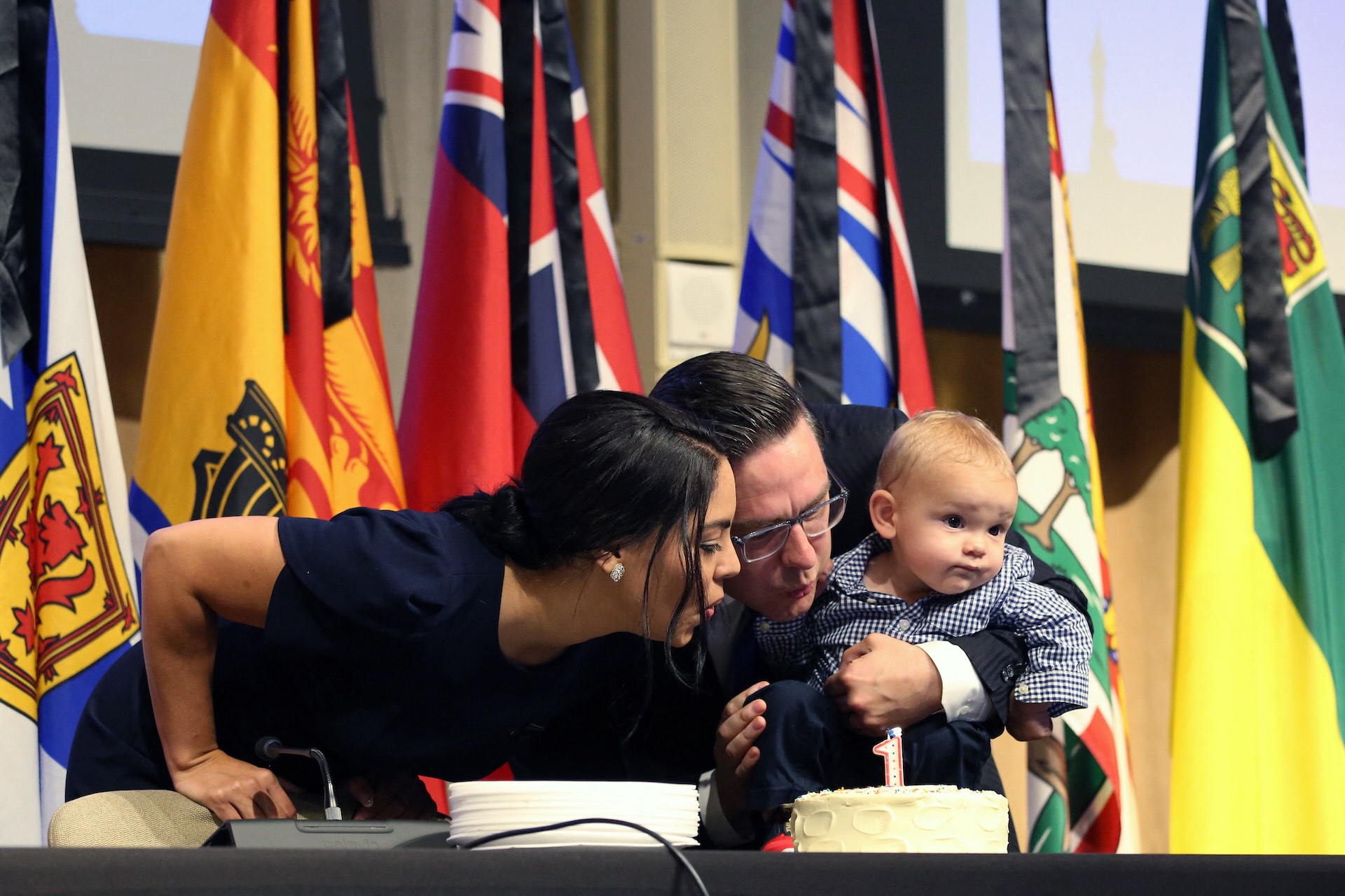 Poilievre holds his one-year-old son and blows out a candle on cake with his wife Anaida. There is a row of provincial flags in the background.