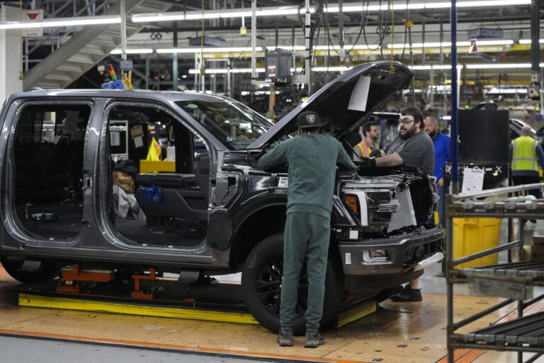 Two employees in a factory work under the hood of a large, grey pickup truck that is partially assembled. The doors and front grill of the truck are not yet attached.