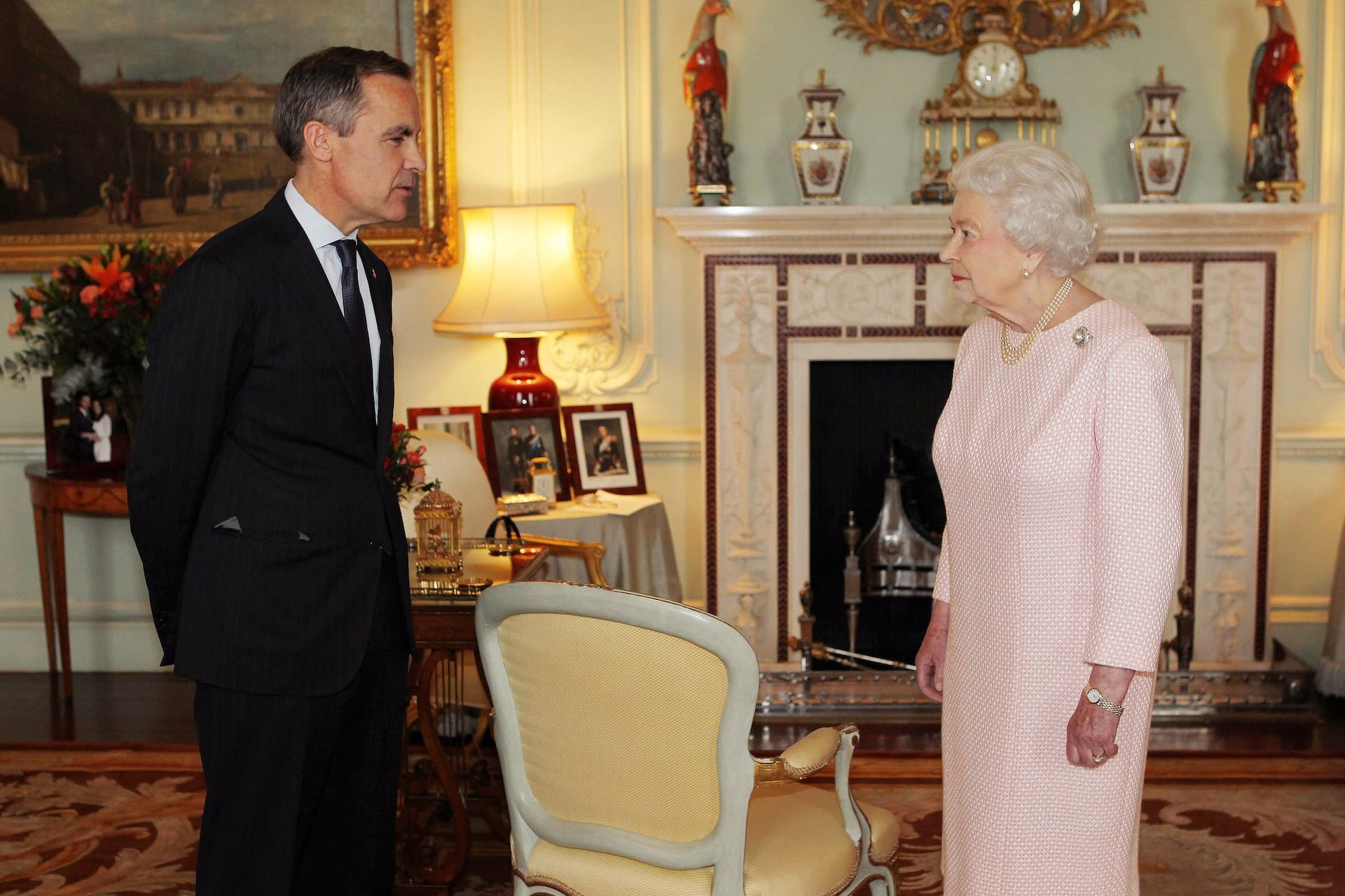 Mark Carney stands in a dark suit in an ornate room in Buckingham Palace a few feet away from Queen Elizabeth II, who is wearing a pink dress and pearls.