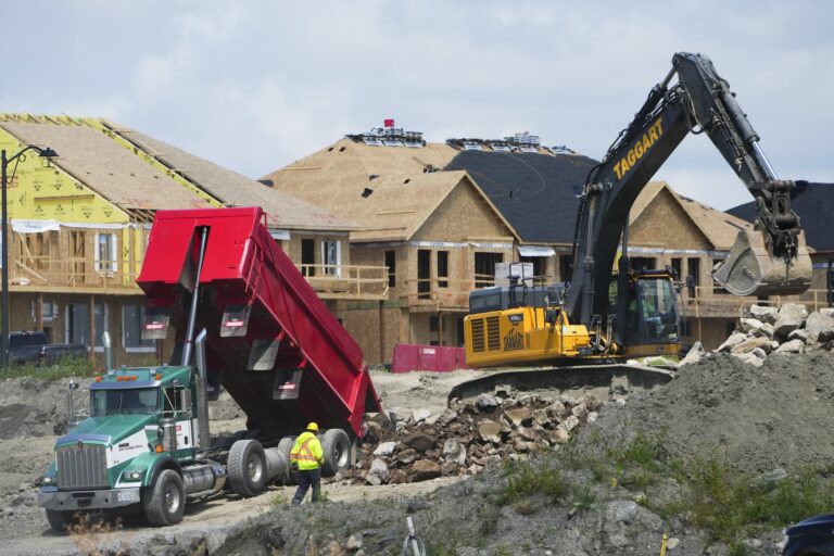 A dump truck and an excavator at work in a construction zone. Half-built homes are behind them.