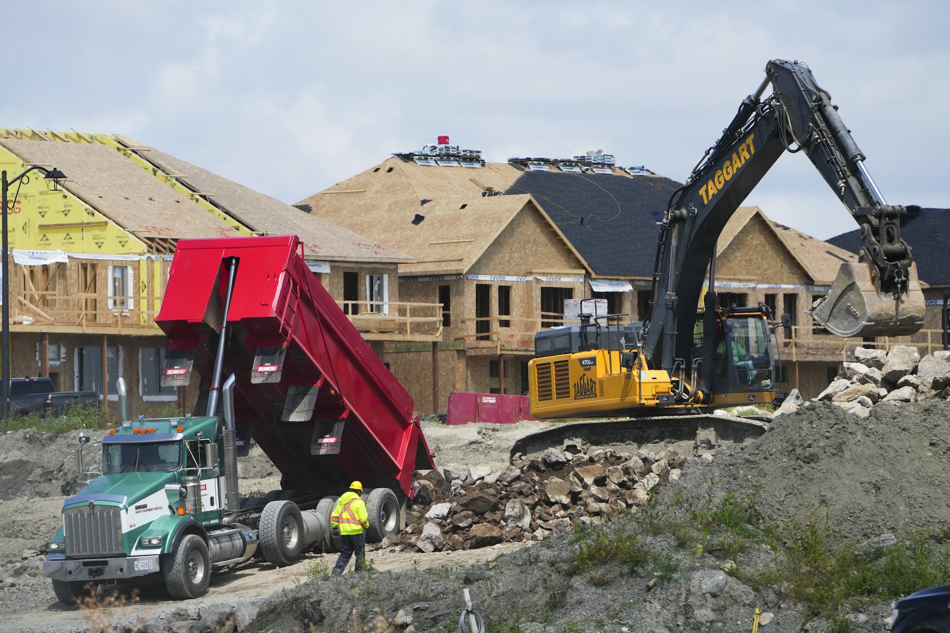 A dump truck and an excavator at work in a construction zone. Half-built homes are behind them.