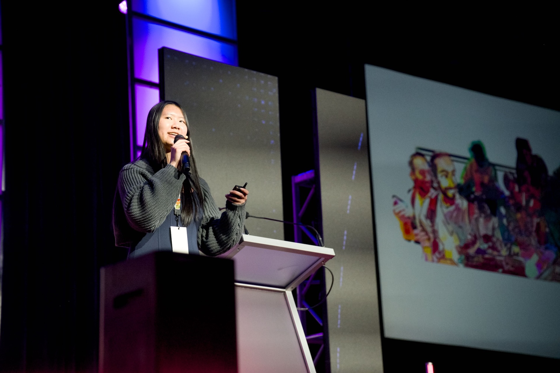 A woman holds a clicker and mic while a screen in the background displays illustrated drawings of people.