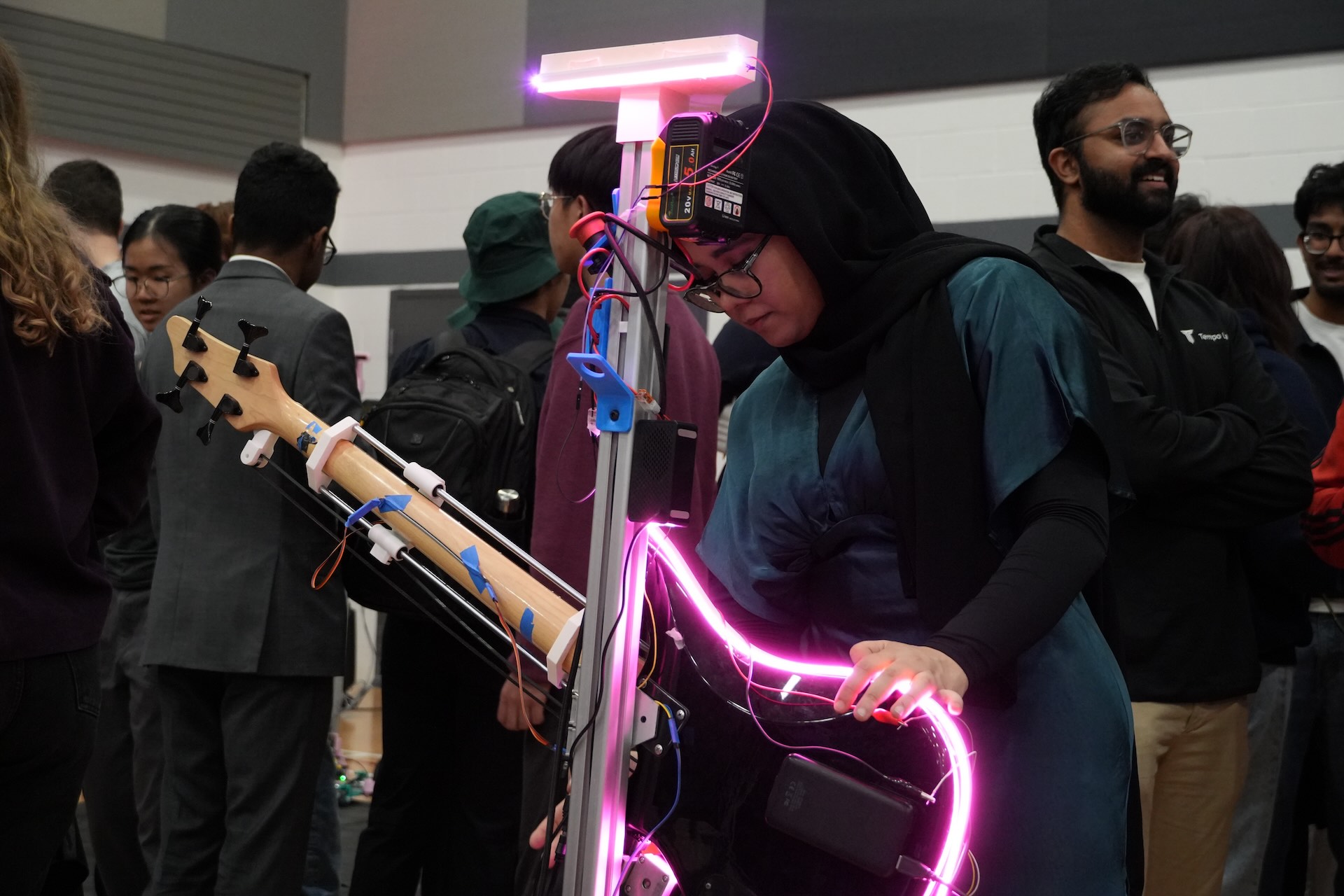 A woman holds a bass lined with pink neon lights that is attached to a stand, with wires connecting it to equipment at the top.