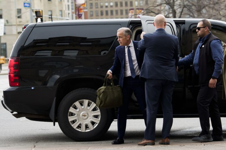 A man in a blue suit holding a green duffel bag steps out of the back door of a large black van while another man holds the door open for him.