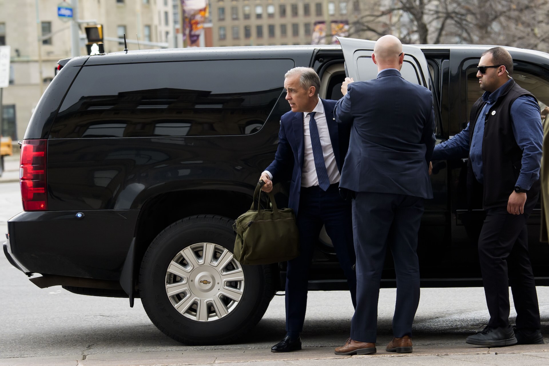 A man in a blue suit holding a green duffel bag steps out of the back door of a large black van while another man holds the door open for him.