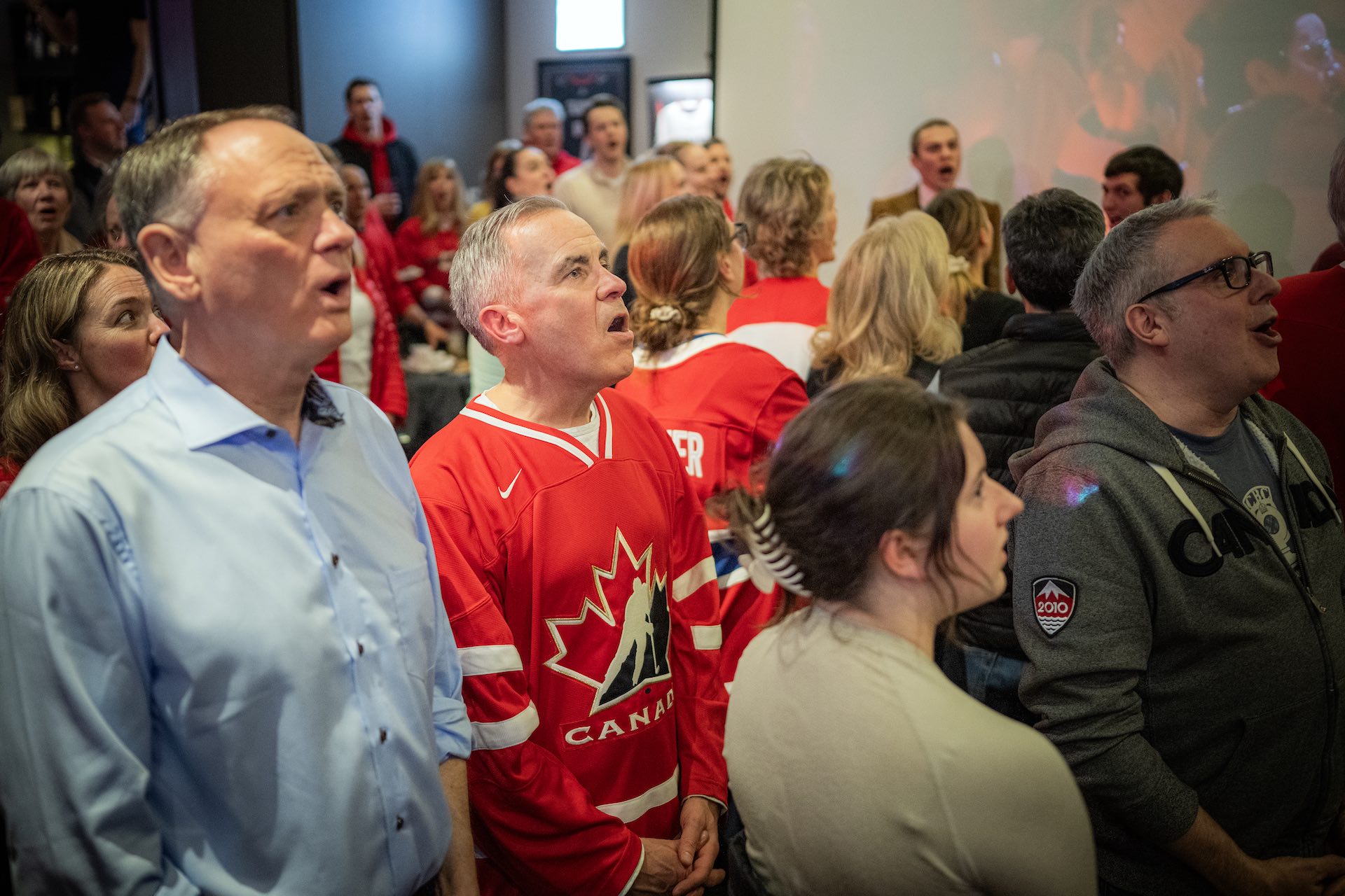 Mark Carney stands in a Team Canada hockey jersey, among a crowd of onlookers in a sports bar, with expression of dismay on his face at what he's just seen on a TV screen.