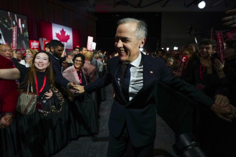 Mark Carney walking down an aisle lined with supporters, touching hands as he passes them. There is a Canadian flag on the wall in the background.