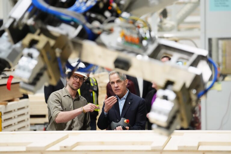 Mark Carney stands next to a worker in a prefab home factory, holding a remote control unit. There is a robotic arm out of focus in the foreground.
