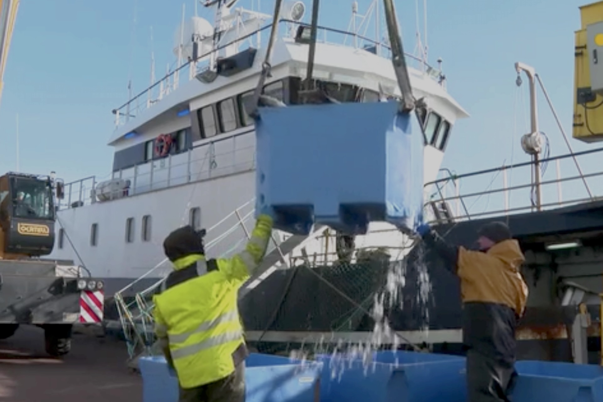 A shot of bins full of fish being lifted by crane onto a dock, with a large fishing boat in the background.