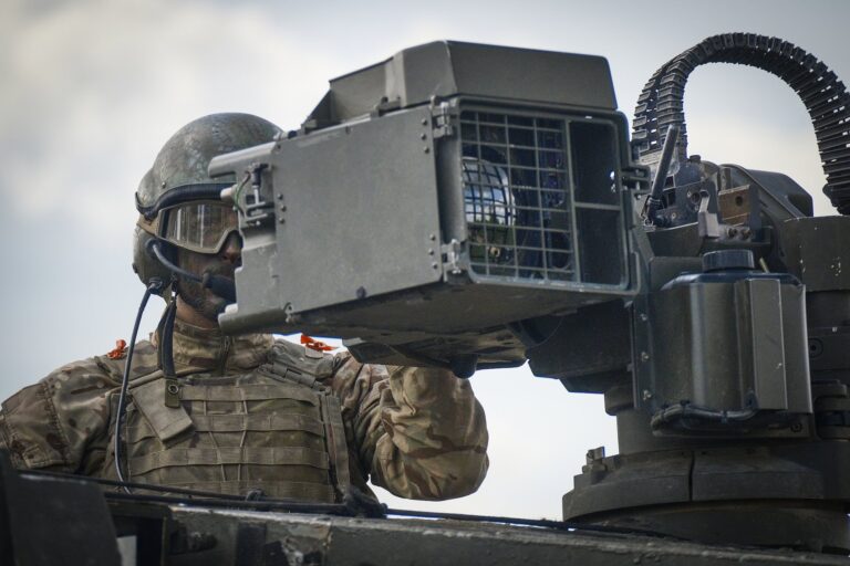 A man in combat gear stands with his chest sticking out the top of a tank. He is looking through equipment connected to a gun turret