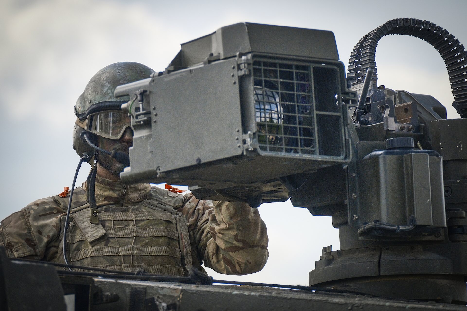 A man in combat gear stands with his chest sticking out the top of a tank. He is looking through equipment connected to a gun turret