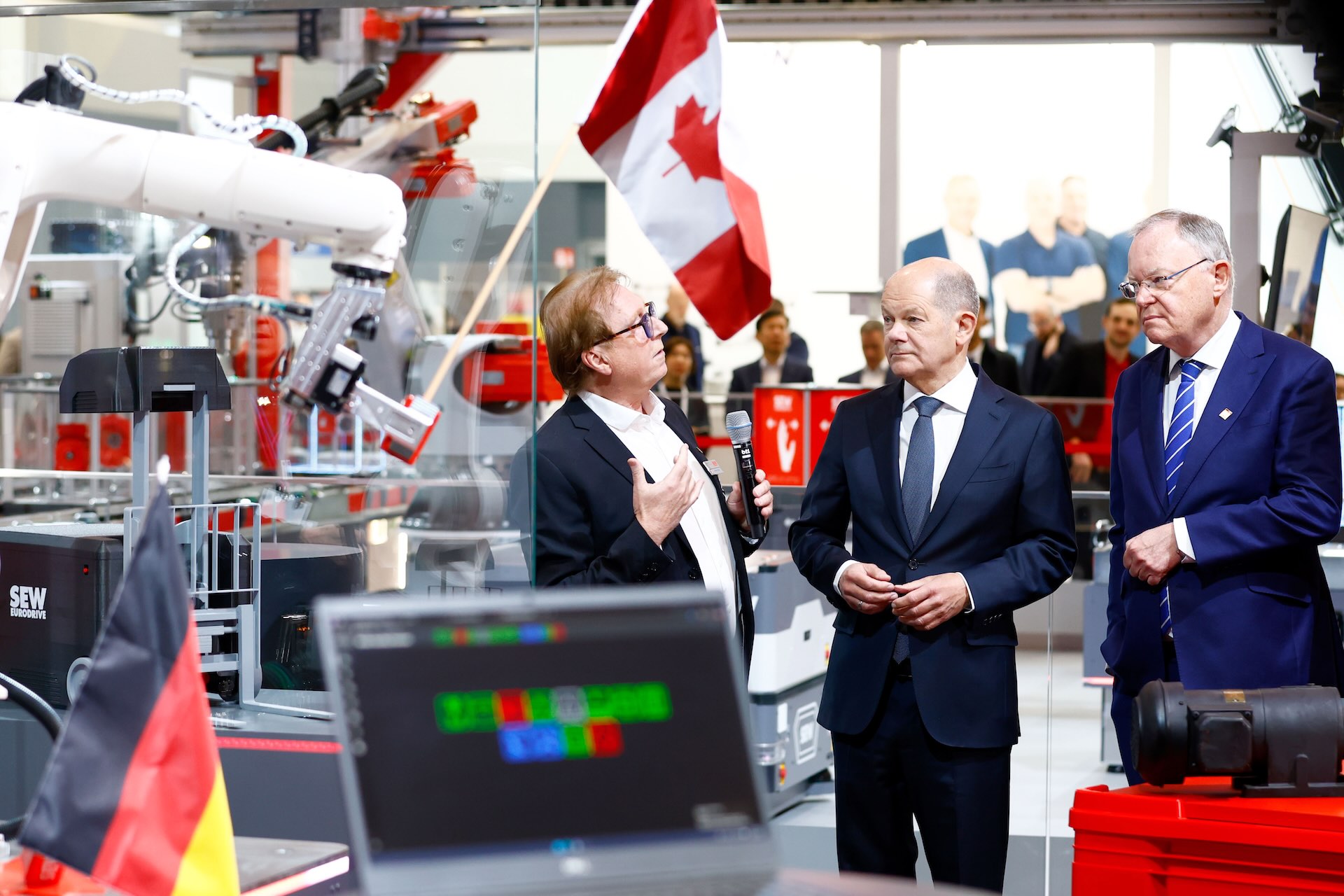 German Chancellor Olaf Scholz talking with two other men in suits at an industrial fair booth with a Canadian flag in the background.