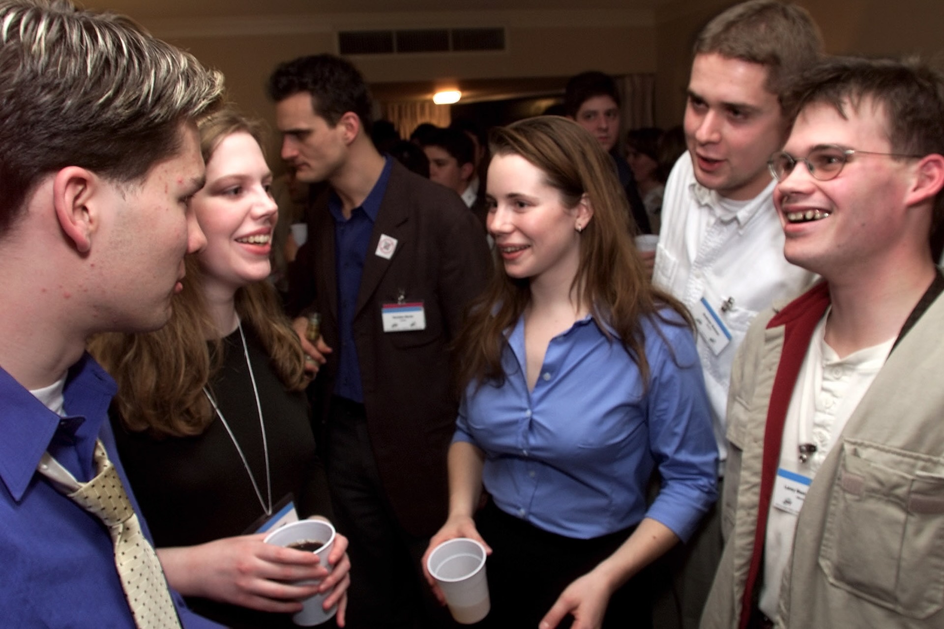 A candid photo of a group of young people, two of whom are holding white plastic drink cups. Pierre Polievre is visible in profile on the far left of the frame.