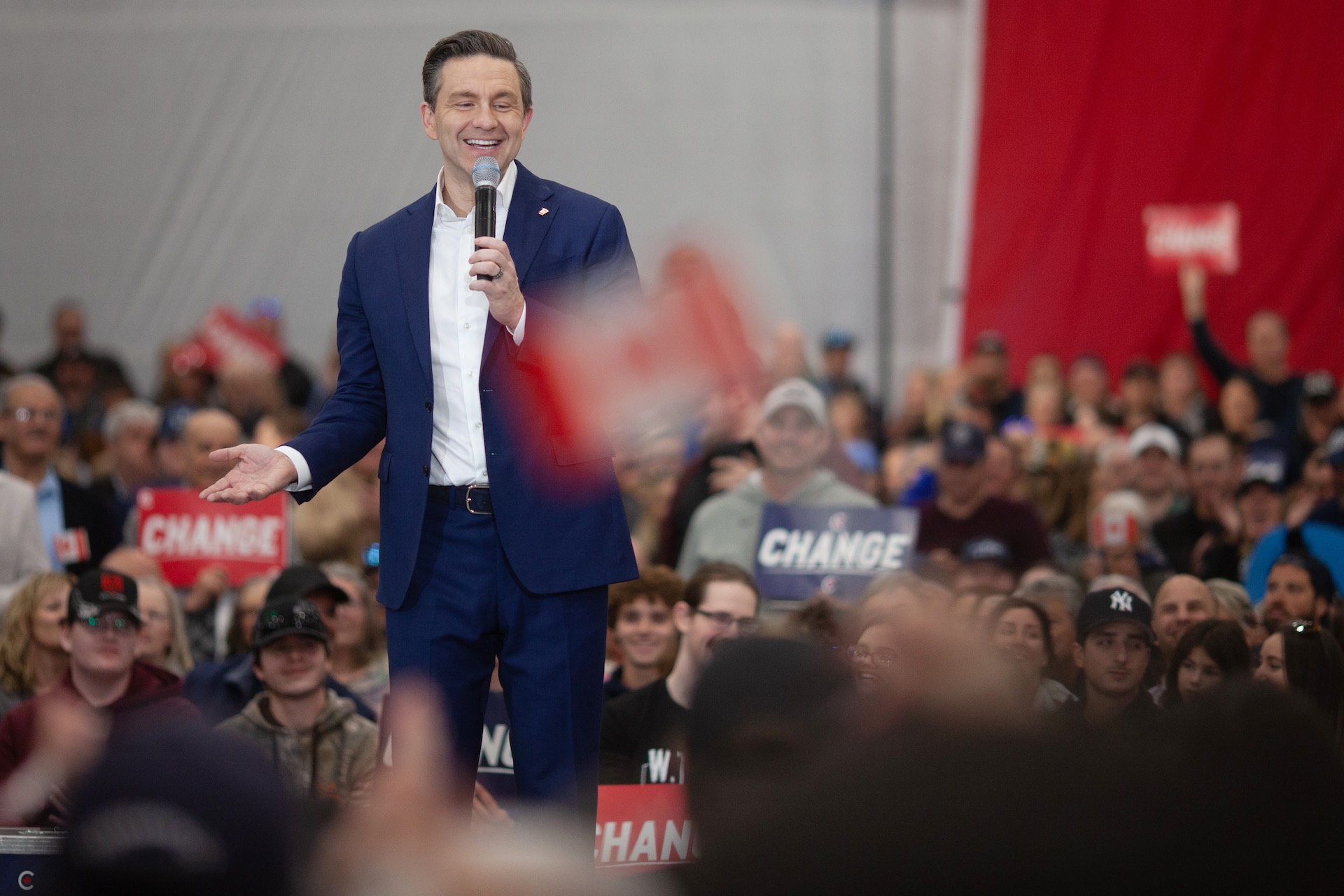 Poilievre smiles while speaking into a microphone on stage; he is surrounded by supporters, some of whom have signs that read "Change." He is wearing a blue suit and an open-necked white shirt.