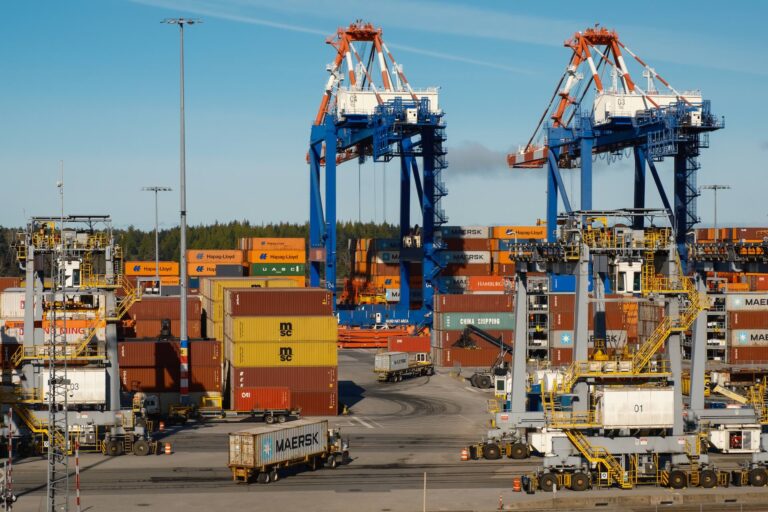Shipping containers stacked at a busy port terminal, with cranes and transport vehicles operating under a clear sky.