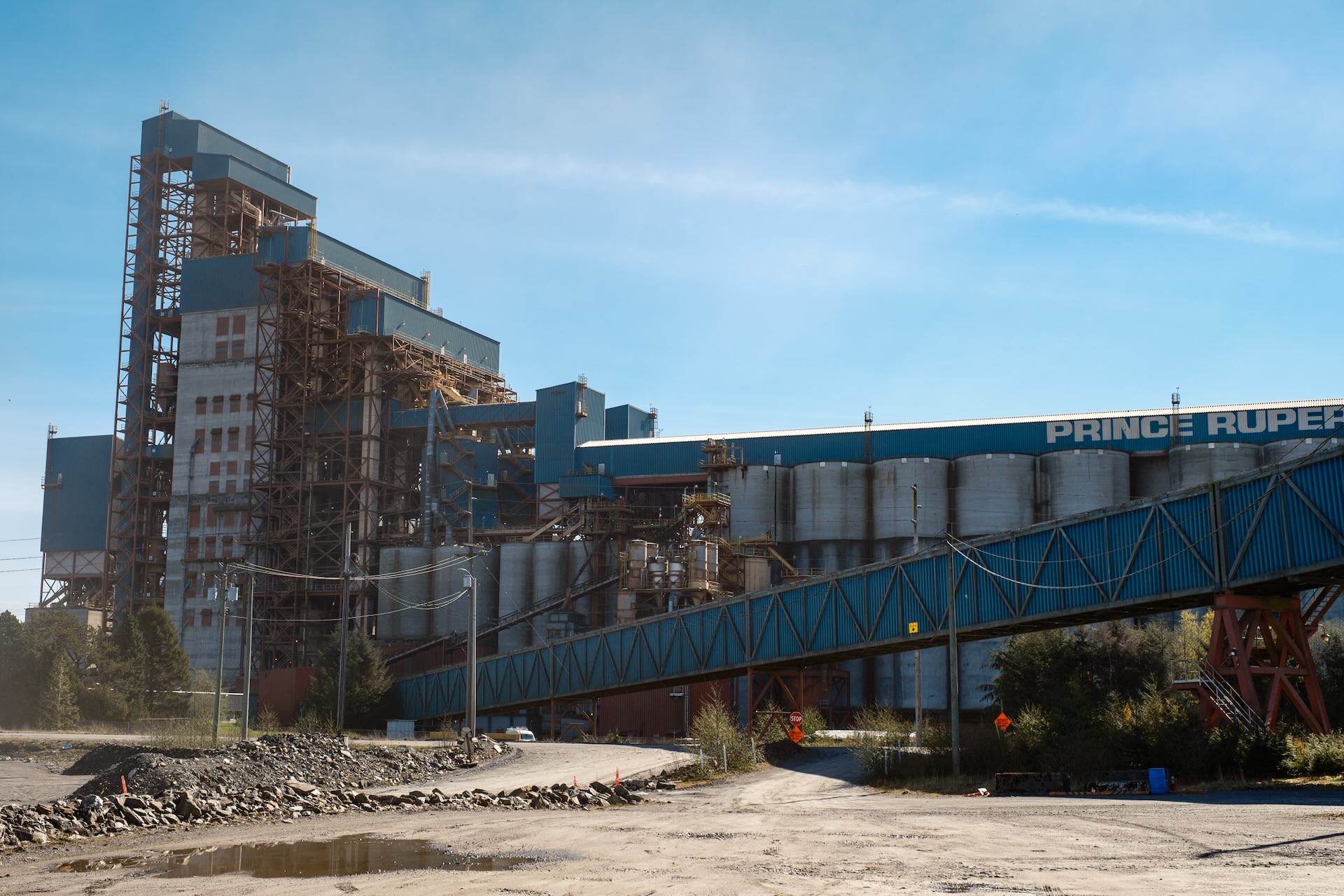A large industrial facility with multiple metal silos, tall structures, and a blue elevated conveyor labeled "Prince Rupert" under a clear sky.