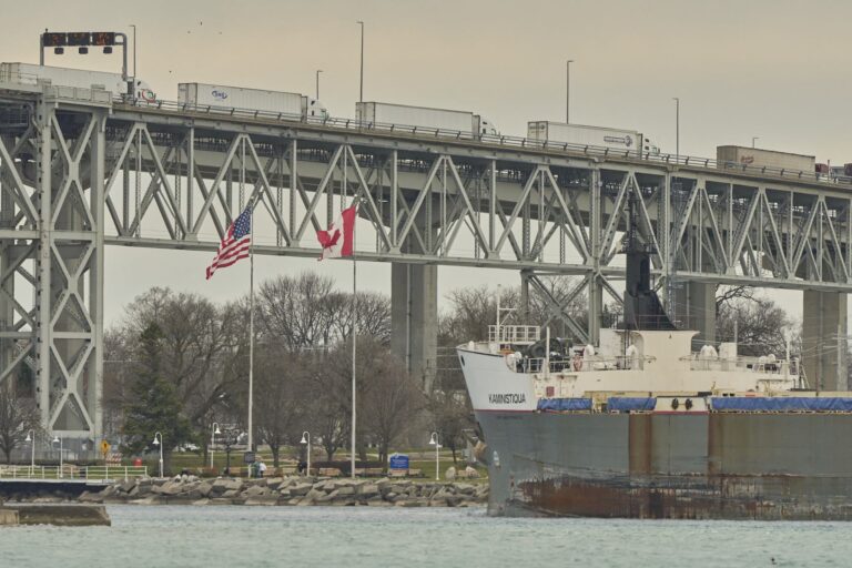 A row of trucks pulling white freight trailers lined up on a bridge. The Canadian and U.S. flags stand on the river shore below the bridge, and the prow of a ship is visible on the right side of the frame.