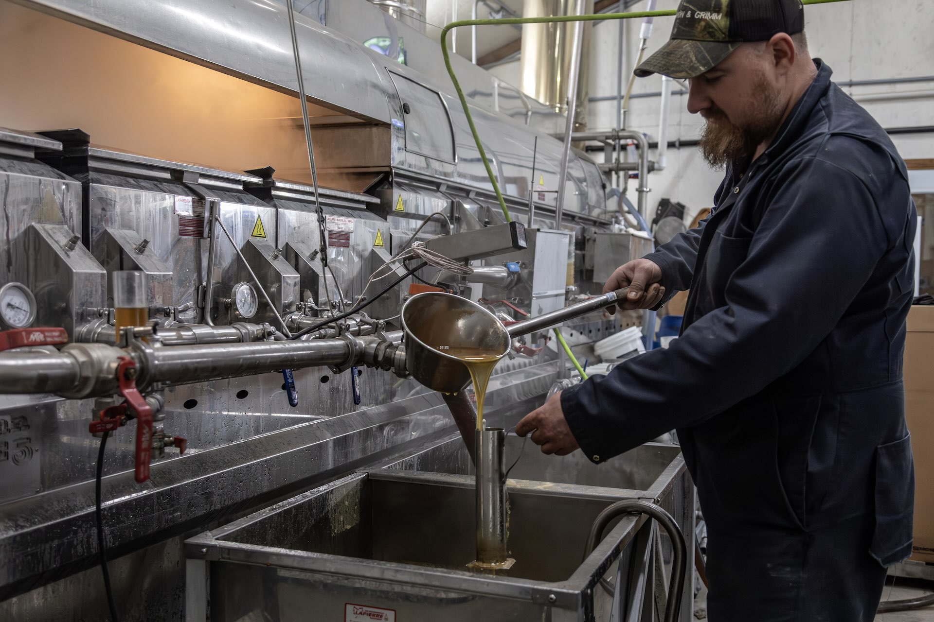 A man in work clothes pours liquid from a ladle into a metal container at an industrial facility with machinery in the background.