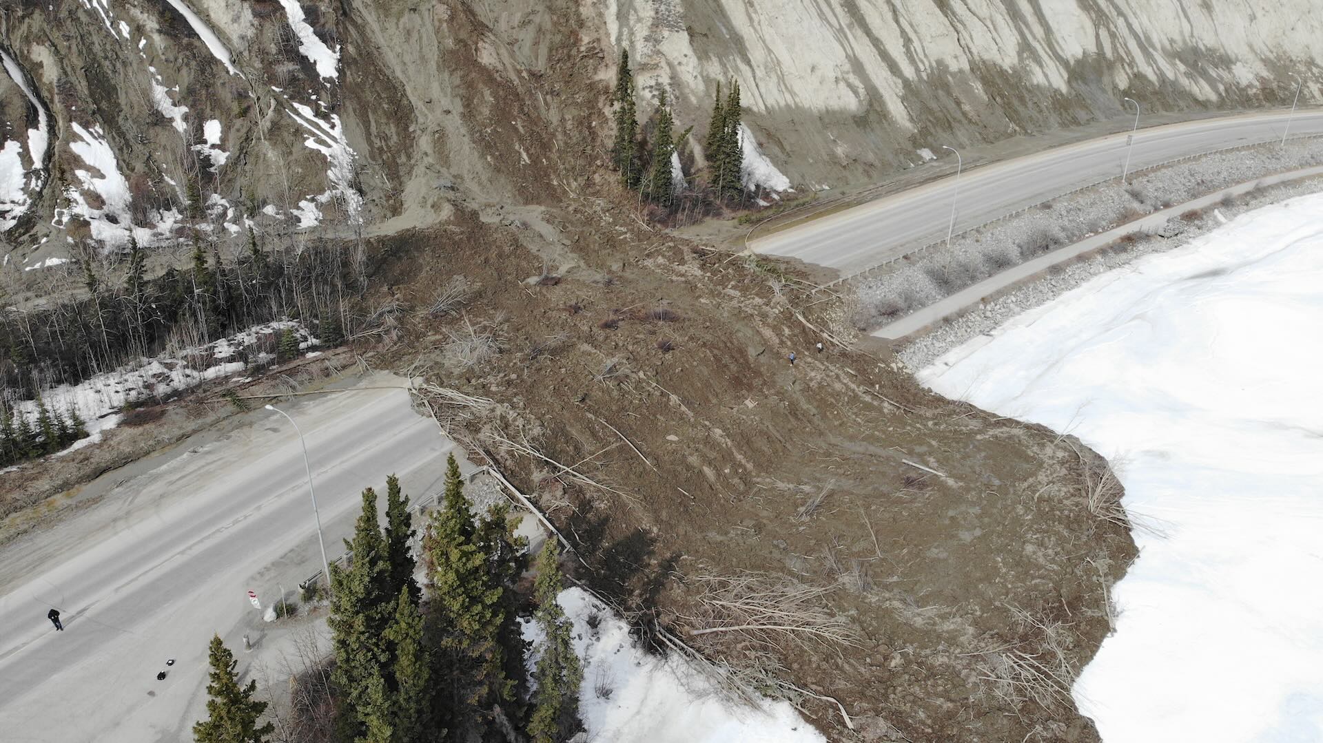 Landslide blocking a road next to a river, with mud and debris covering the roadway. Snow is visible on the ground, and trees are on the sides.