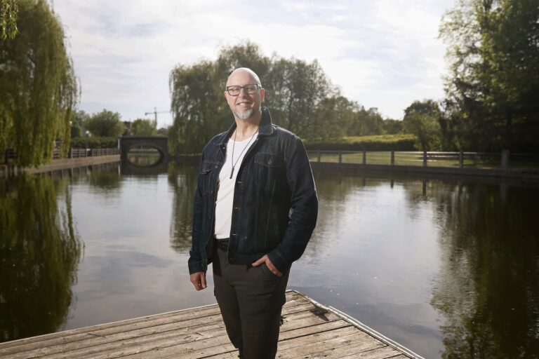 A man wearing glasses, a white t-shirt, and a denim jacket stands on a wooden dock by a calm pond with trees and a bridge in the background.