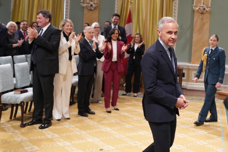 A shot of Mark Carney glancing at the camera as he crosses the yellow carpet of Rideau Hall. Liberal cabinet members Anita Anand, François-Philippe Champagne, Melanie Joly and Dominic LeBlanc are visible in the background.