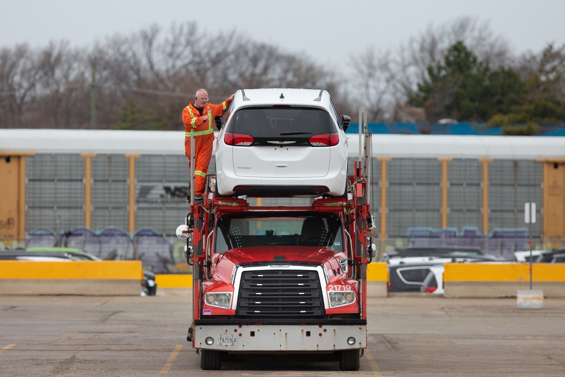 A worker in an orange safety suit secures a white van on the upper deck of a red car transport truck in an industrial parking lot. Trees and train cars are in the background.