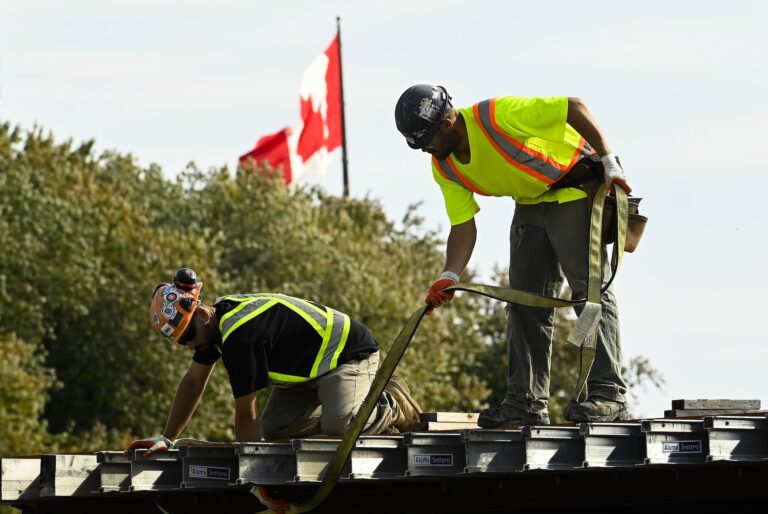 A shot of two construction workers in yellow reflective workwear on top of a structure. There are trees and a Canadian flag in the background.