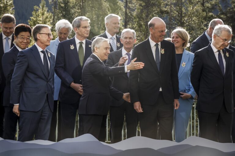 A shot of François-Philippe Champagne standing on a dais among besuited fellow finance ministers. He is gesturing with one hand, as if to lead the group off the stage. There are tall evergreen trees in the background.