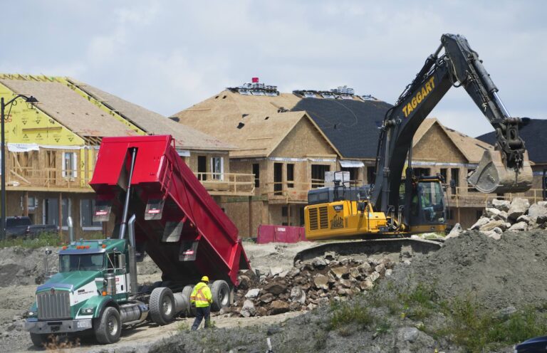 A dump truck unloads rubble while an excavator moves rocks at a construction site with partially built houses in the background.