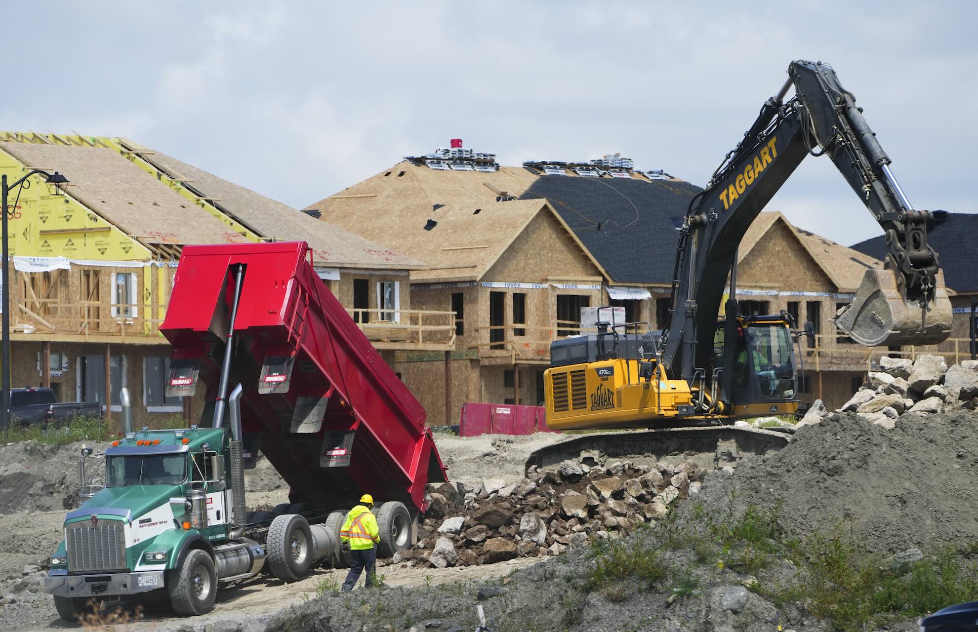 A dump truck unloads rubble while an excavator moves rocks at a construction site with partially built houses in the background.