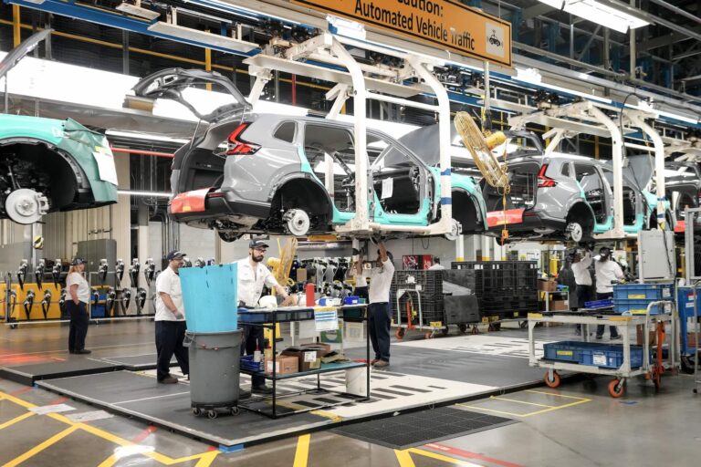 Workers assemble vehicles on an automated production line in a factory, with car frames suspended above work stations and equipment nearby.