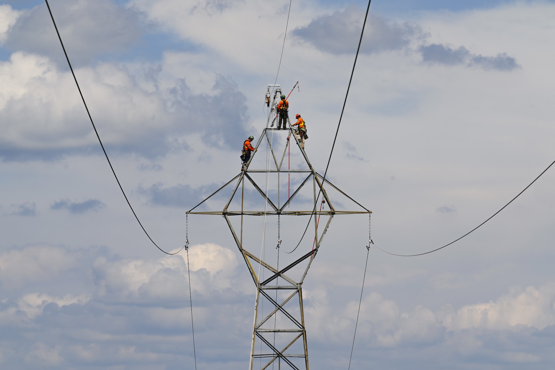 Three workers in safety gear perform maintenance on the top of a metal electrical transmission tower against a cloudy sky.