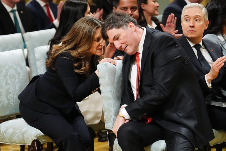 A candid shot of Chrystia Freeland leaning forward from the second row of a cabinet swearing-in ceremony to speak into Dominic LeBlanc's ear. LeBlanc has his head tilted close to hers. Francois-Philippe Champagne is looking on.
