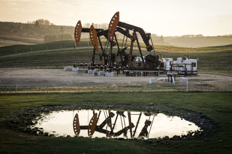 Several oil pumpjacks operate on a grassy field, with their reflections visible in a nearby pond under a cloudy sky.