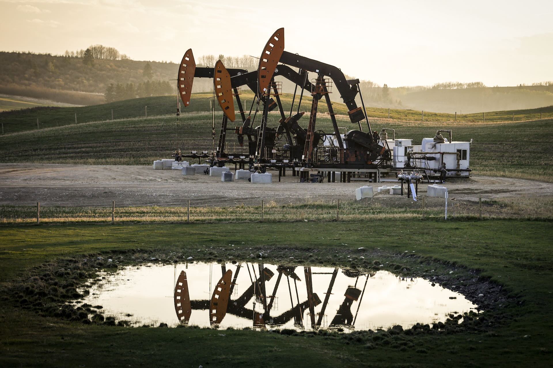 Several oil pumpjacks operate on a grassy field, with their reflections visible in a nearby pond under a cloudy sky.