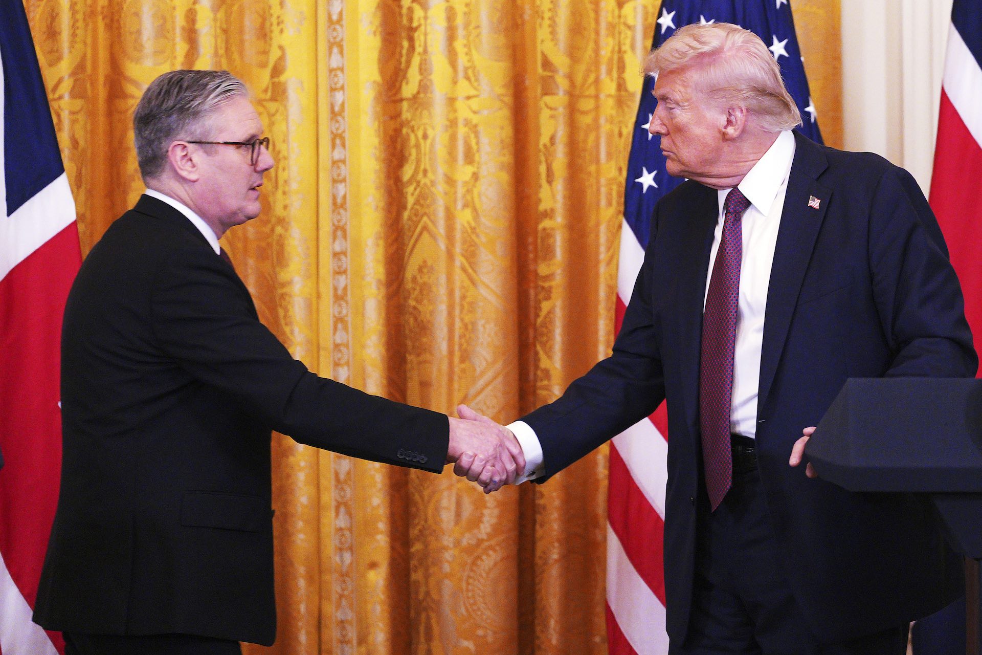 Keir Starmer shaking hands with Donald Trump with a golden curtain and U.S. and U.K. flags in the background.