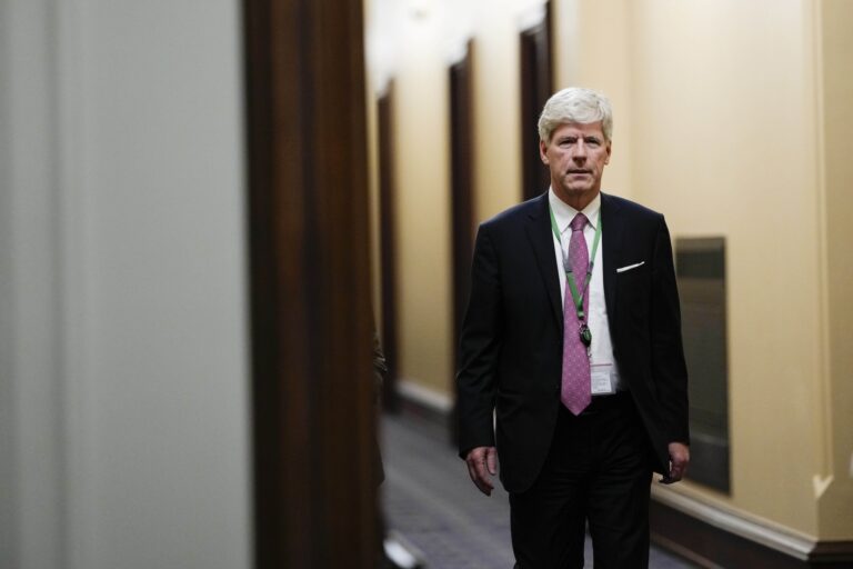 A head-on shot of Tim Hodgson walking down a corridor as he looks directly at the camera. He's wearing a dark suit and a fuschia tie. Hodgson is tall and has thick, white hair.