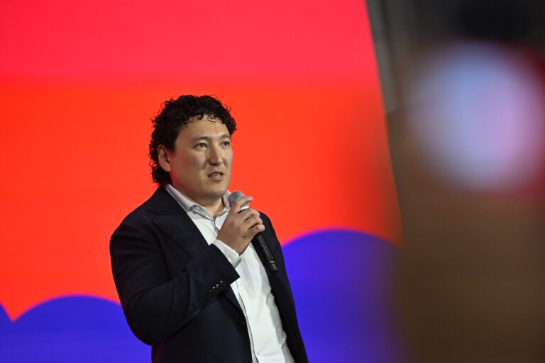 A man in a suit jacket holding a microphone stands in front of a multicoloured backdrop.